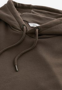 Sweat-shirt à capuche marron avec des cordons épais et des œillets métalliques, montrant une étiquette blanche partiellement visible à l'intérieur du col.