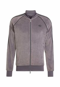 Chaqueta morada con cremallera, fabricada en un tejido suave, con franjas laterales negras, puños acanalados y un pequeño logotipo en el pecho.