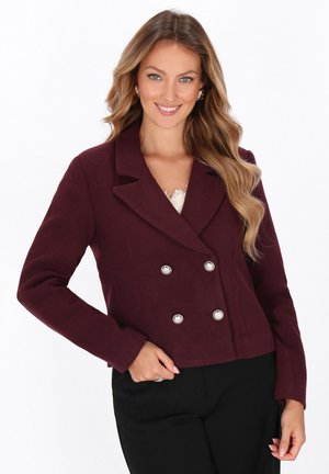 Donna che sorride, con indosso un blazer doppiopetto bordeaux con bottoni argentati, pantaloni neri e capelli biondi ondulati su sfondo bianco.