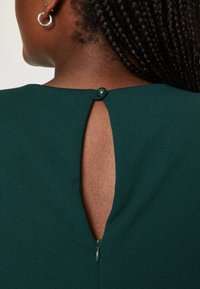 Donkergroene jurk met een ronde halslijn, met een keyhole uitsnijding op de rug, vastgezet met een bijpassende stoffen knop en ritsdetail.