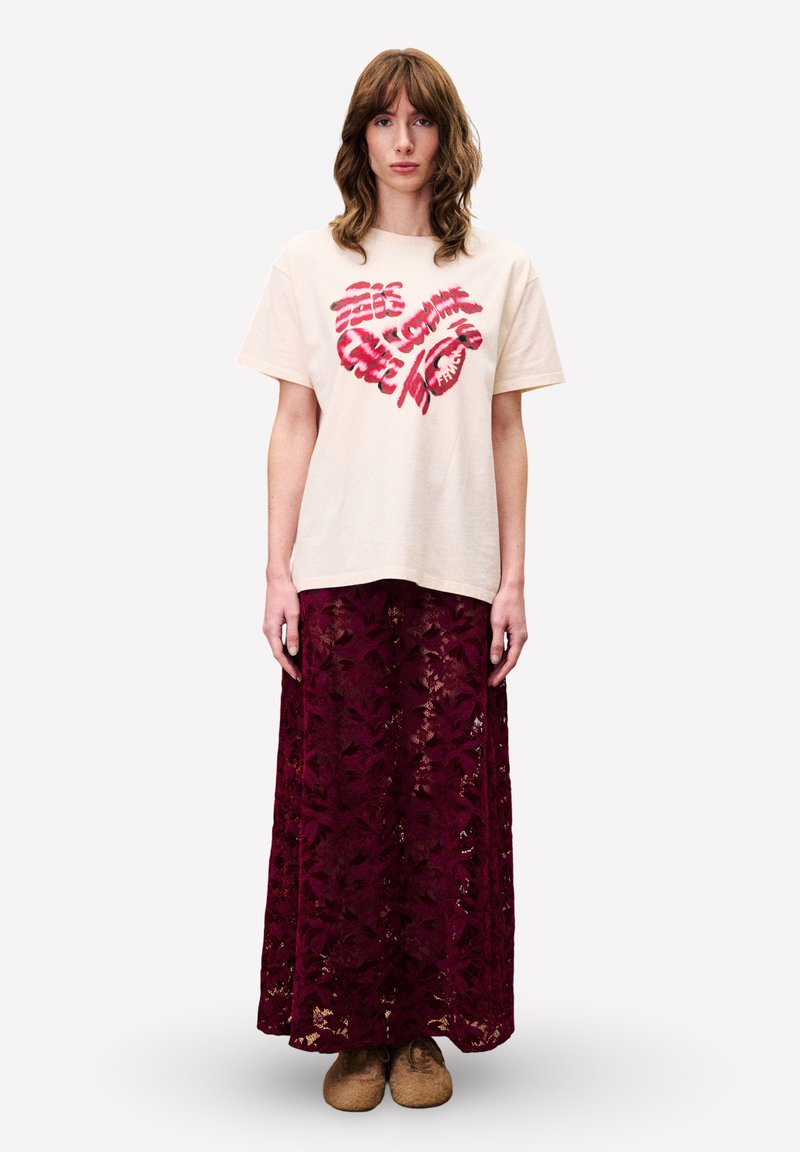 Cremfarbendes Baumwoll-T-Shirt mit rotem Grafikdesign und einem tiefburgunderfarbenen Spitzen-Maxirock. Das Modell trägt beiges Schuhwerk und steht vor einem schlichten Hintergrund.