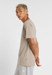Camiseta de manga corta beige hecha de tela suave con cuello redondo. Presenta una pequeña etiqueta con el logotipo en el dobladillo. Modelo vistiendo pantalones de chándal grises.