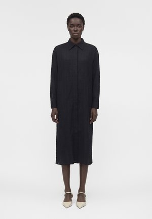 Γυναίκα που στέκεται, φοράει μακρύ μαύρο φόρεμα με κουμπιά, γιακά και μακριά μανίκια, συνδυασμένο με λευκά μυτερά τακούνια με λουράκια στον αστράγαλο.