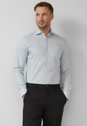 Mann trägt ein hellblau gestreiftes Hemd, das in schwarze Hosen gesteckt ist, steht mit den Händen in den Taschen vor einem schlichten Hintergrund.