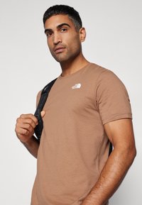 Camiseta de algodón marrón con mangas cortas y cuello redondo; presenta un logo blanco en el pecho; la persona sostiene una correa de bolsa negra.