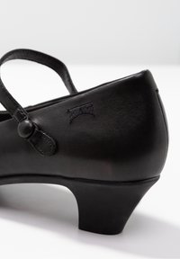 Chaussure Mary Jane en cuir noir avec petit talon et bride boutonnée sur fond blanc uni.