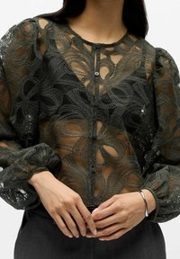 Sheer lace blouse in dark green with intricate floral pattern, round neckline, six button closure, and puffed sleeves. Textured fabric detail.