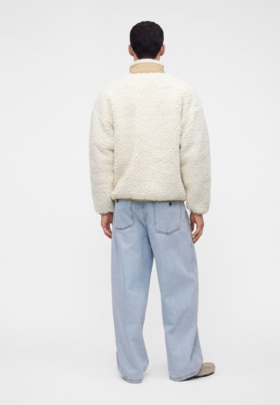 Άνδρας που φοράει μπεζ φλις μπουφάν και ανοιχτά μπλε φαρδιά τζιν, στεκόμενος με την πλάτη σε απλό λευκό φόντο.