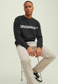 Schwarzer Sweatshirt mit weißem "ORIGINALS™"-Text, kombiniert mit hellbeigen Hosen und schwarz-weißen Sneakern. Das Model sitzt auf einem Hocker.