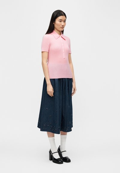 Γυναίκα με μακριά σκούρα μαλλιά, ντυμένη με ροζ πόλο με κοντά μανίκια και κουμπιά, μπλε σκούρα μίντι φούστα με κεντήματα λουλουδιών, λευκές κάλτσες και μαύρα παπούτσια στιλ Mary Jane με τακούνι.