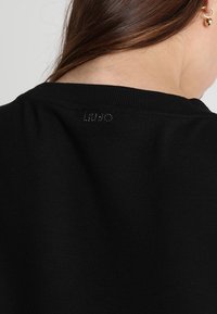 Svart tröja med rund halsringning och ribbade muddar. Har en subtil logotypdetalj på baksidan av nacken. Mjuk textur, figursydd form.