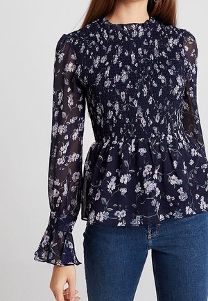 Femme portant un chemisier bleu marine à manches longues avec imprimé floral blanc, corsage smocké, encolure volantée et manches évasées, associé à un jean bleu.