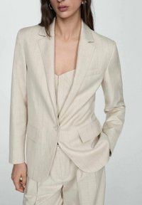 Blazer beige clair ajusté avec un bouton unique, des revers crantés et de fines rayures verticales. Confectionné dans un tissu lisse avec une légère brillance.