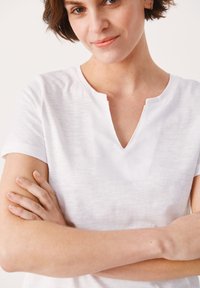 T-shirt bianco a maniche corte con scollo a V, realizzato in un tessuto leggero e texturizzato. Il colletto presenta un sottile dettaglio a intaglio. Le braccia sono incrociate sul petto.