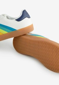 Sneaker blanche avec un talon bleu, ornée de rayures multicolores en bleu, jaune et vert. Semelle extérieure en gomme texturée visible.