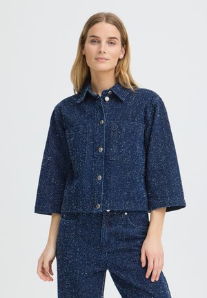 Jeune femme portant une veste en denim bleu foncé mouchetée avec des boutons et un jean taille haute assorti, debout devant un fond uni.