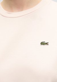 Gros plan d’un t-shirt beige clair à col rond avec un petit logo crocodile vert brodé sur le côté gauche de la poitrine.