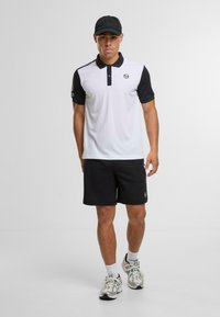 Herrpolo i vitt och svart med korta ärmar, en knäppt krage och en logodetalj. Matchas med svarta shorts och sportskor.