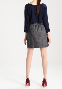 Žena stojící, oblečená v námořnické blůze, šedé sukni ke kolenům a bordó vysokých podpatcích s červenými podrážkami, stojí zády k prostému pozadí.