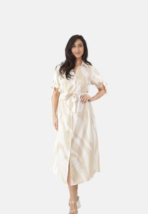 Mujer con cabello largo y oscuro que lleva un vestido camisa midi beige y blanco con estampado, mangas atadas y sandalias transparentes de tacón, de pie con la mano en la cadera.