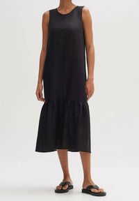Svart ärmlös klänning med avslappnad passform, med en flerskikts nederdel och tillverkad av ett lätt material. Matchas med svarta platta sandaler.