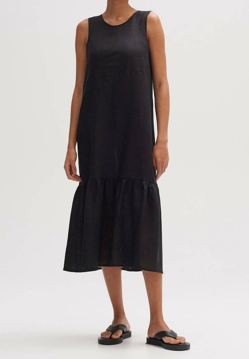 Svart ärmlös klänning med avslappnad passform, med en flerskikts nederdel och tillverkad av ett lätt material. Matchas med svarta platta sandaler.