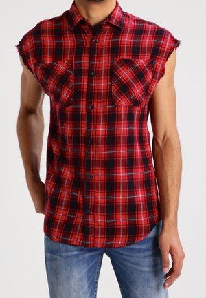Homme portant une chemise à carreaux rouge et noire sans manches avec deux poches poitrine et un jean bleu sur un fond uni.
