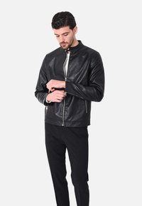 Giacca di pelle nera con colletto rotondo, cerniera frontale e tasche con cerniera. Indossata sopra una camicia bianca e abbinata a pantaloni neri.