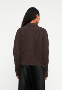 Γυναίκα με μακριά κυματιστά μαλλιά, φορούσα ένα σκούρο καφέ φλις πουλόβερ και μια μαύρη πλισέ φούστα, απεικονιζόμενη από πίσω σε λευκό φόντο.