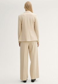 Traje beige a medida que presenta una chaqueta ajustada y pantalones de pierna ancha, confeccionado con un tejido suave y con una textura sutil. Zapatos negros completan el conjunto.