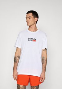 Man with tattoos wearing white "Berlin Run" T-shirt and bright orange shorts looking to the side against a plain background.