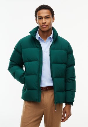 Hombre que lleva una chaqueta acolchada verde oscuro sobre una camisa azul clara y pantalones marrones, de pie con una mano en el bolsillo.