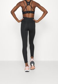 Leggings sportivi neri con vita alta, abbinati a un reggiseno sportivo nero con spalline e dettagli con logo. Modella che indossa scarpe da corsa rosa chiaro.