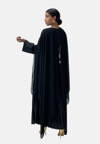 Czarna sukienka z szyfonu o zwiewnym kroju, długimi rękawami i warstwowym drapowaniem. Złote akcenty i plisy przy dolnej krawędzi.