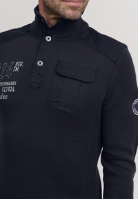 Tengerészkék pulóver magas gallérral, gombos nyakkal és mellzsebbel. Texturált kötött anyag és kontrasztos vállpanelek.