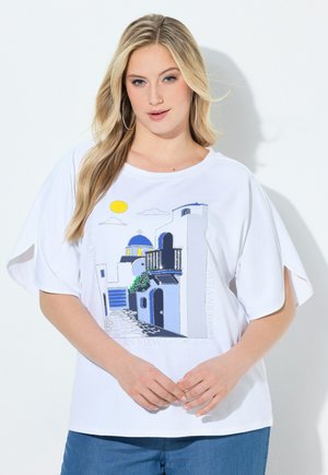 Lyshåret kvinde iført hvid T-shirt med blå og gul illustration af en bygning i Santorini-stil, kombineret med blå jeans, stående mod en hvid baggrund.