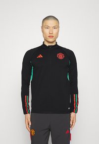 Camiseta deportiva negra de manga larga con paneles laterales verdes. Presenta acentos rojos, un cuello alto y un logotipo del Manchester United en el pecho.