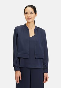 Veste cropped bleu marine avec une finition satinée lisse, col mandarin, poignets élastiqués et design à ouverture frontale. Associée à un haut assorti.