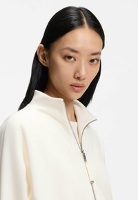 Weiße Jacke mit Reißverschluss und hohem Kragen, glatter Textur und silbernen Reißverschlussdetails. Das Modell hat glattes schwarzes Haar und ein neutrales Make-up.