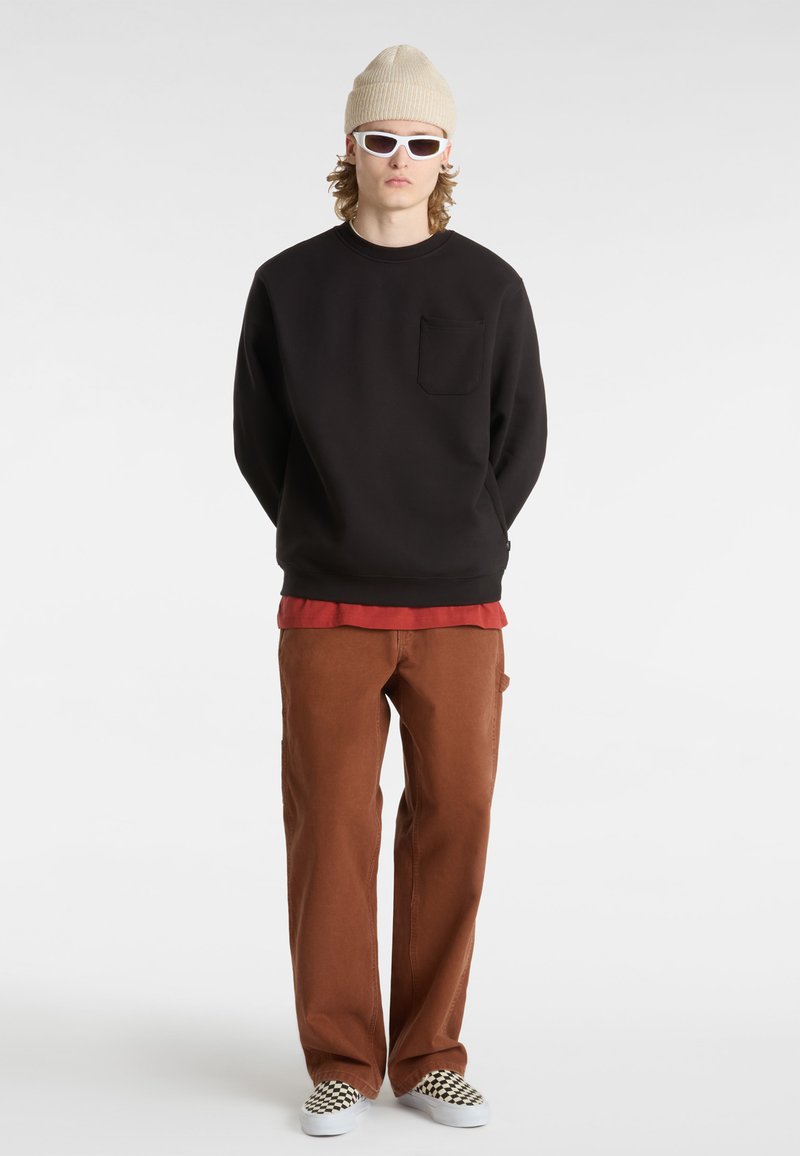 Zwarte sweatshirt met zak, rode gelaagde shirt, bruine broek en geruite instap schoenen. Beige gebreide beanie en witte zonnebril.