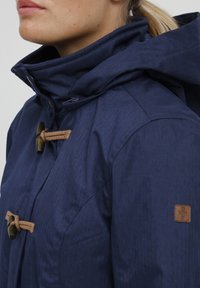 Mörkblå jacka med en texturerad, fiskbensmönster. Har en huva, snoddknäppning och en brun logotagg på ärmen.