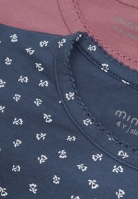 Două tricouri pentru copii așezate unul peste altul. Unul este bordeau, celălalt este de culoare bleumarin cu motive florale albe. Ambele au guler din dantelă și etichete.