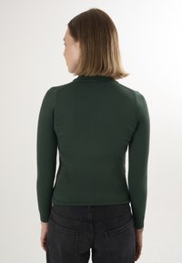 Mørkegrøn tætsiddende langærmet rullekravetrøje med ribbet struktur og strømlinet design, kombineret med sorte højtaljede bukser.