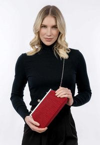 Clutch plissettata rossa con hardware argentato e catena a spalla, tenuta da una persona che indossa un dolcevita a coste nero e pantaloni neri.