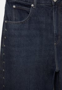 Jean en denim bleu foncé avec poche avant, rivets en métal et une rangée verticale de petits clous en forme d'étoile le long de la couture latérale gauche.