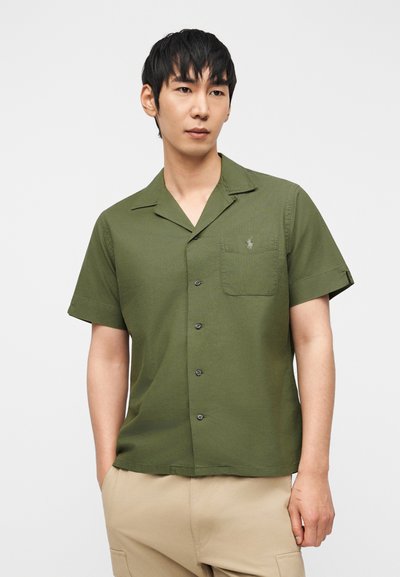 Άνδρας που φοράει πουκάμισο με κουμπιά, κοντά μανίκια σε ελιά πράσινο χρώμα με τσέπη στο στήθος και μπεζ παντελόνι, στέκεται μπροστά σε απλό ανοιχτό φόντο.