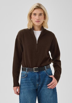 Femme blonde portant un pull zippé marron foncé, un sous-vêtement blanc, un jean bleu, une ceinture noire et des boucles d'oreilles en perles, debout avec une main dans la poche.