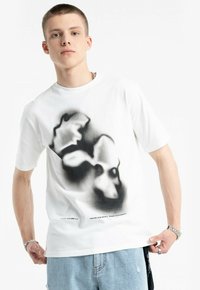 Biała bawełniana koszulka z krótkim rękawem, z klasycznym okrągłym dekoltem, z czarnym abstrakcyjnym wzorem graficznym z przodu. Zauważalny szczegół tekstowy na dole.