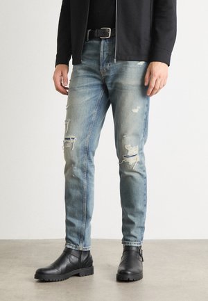 Mies pukeutuneena vaaleansinisiin kuluneisiin farkkuihin, mustaan vyöhön, mustaan takkiin ja mustiin nahkasaappaisiin, seisomassa harmaalla lattialla.