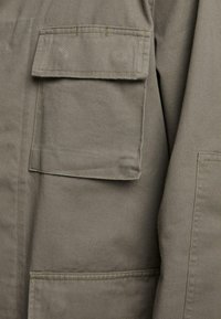 Chaqueta verde oliva con dos grandes bolsillos con solapa, detalles cosidos y un tejido de algodón texturizado. Diseño minimalista, sin cierres visibles.